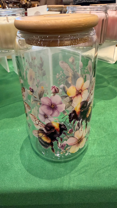 Clear glass jar with wooden lid featuring floral and bee design on a green surface.
