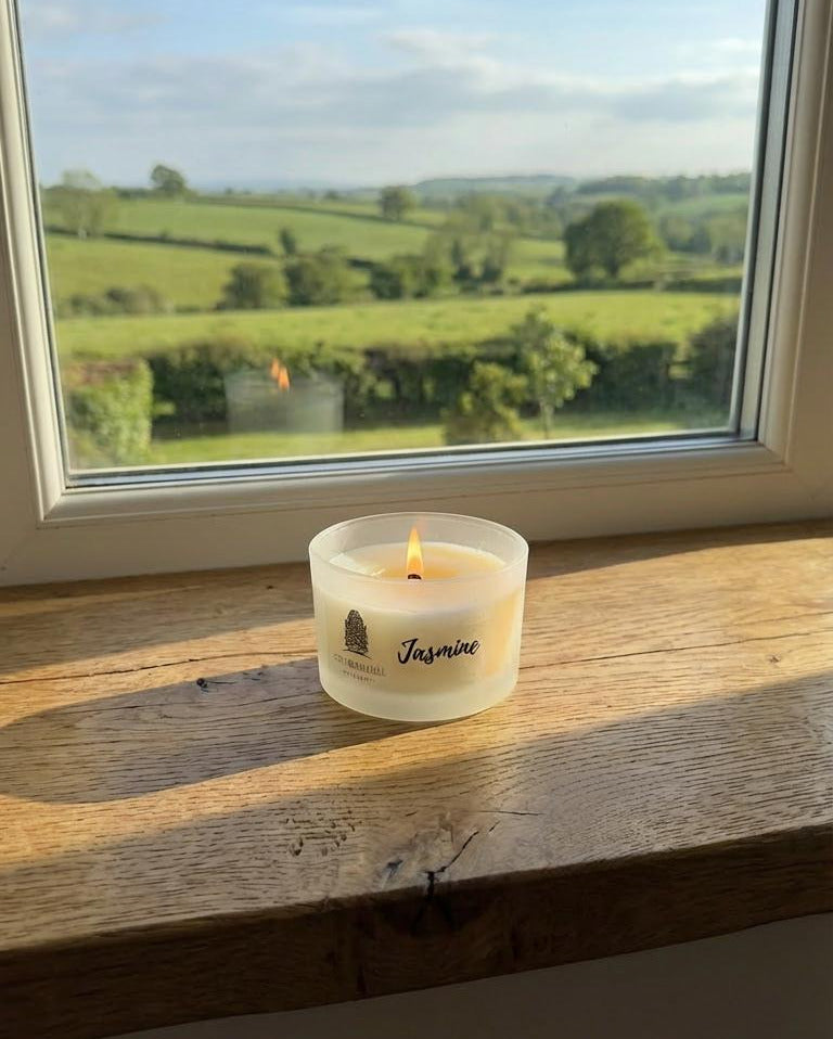 Candle labeled 'Jasmine' on a windowsill with a view of green fields and trees.
